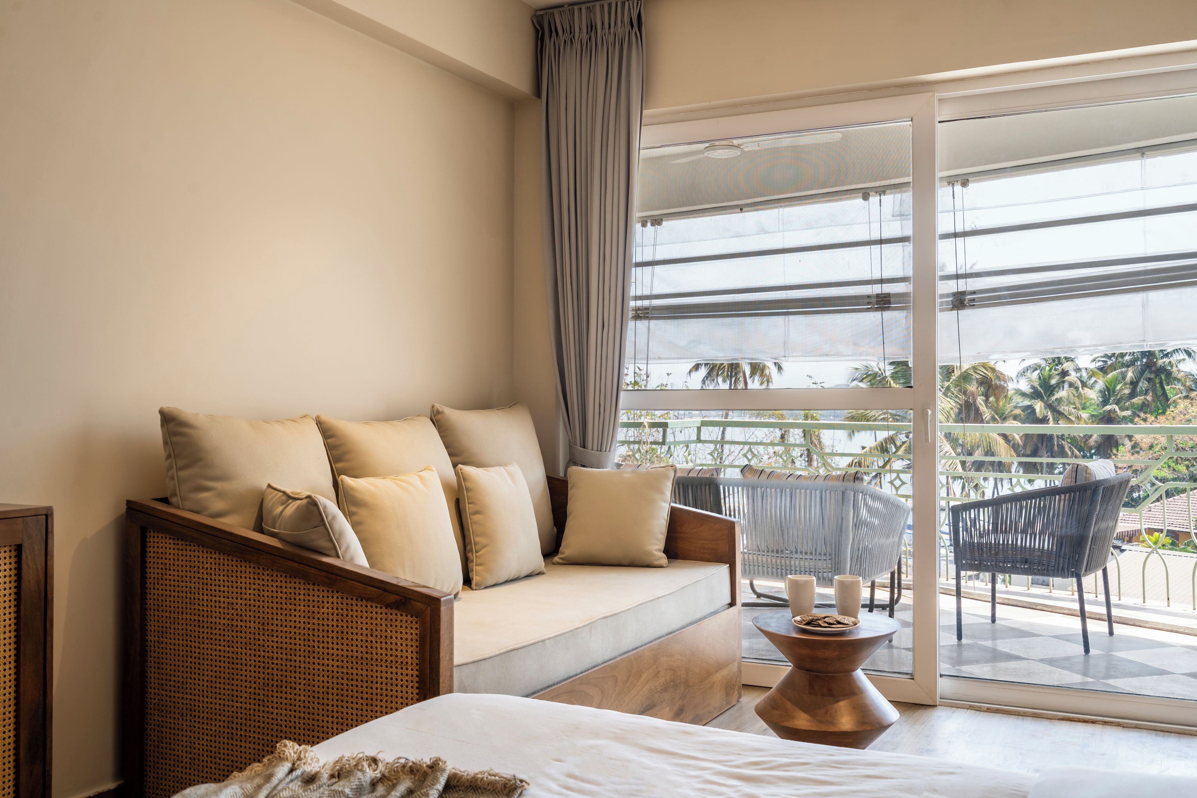 Ein Zimmer mit einer Couch, einem Bett, einem Tisch mit Kerzen und Blick auf Palmen durch große Fenster.