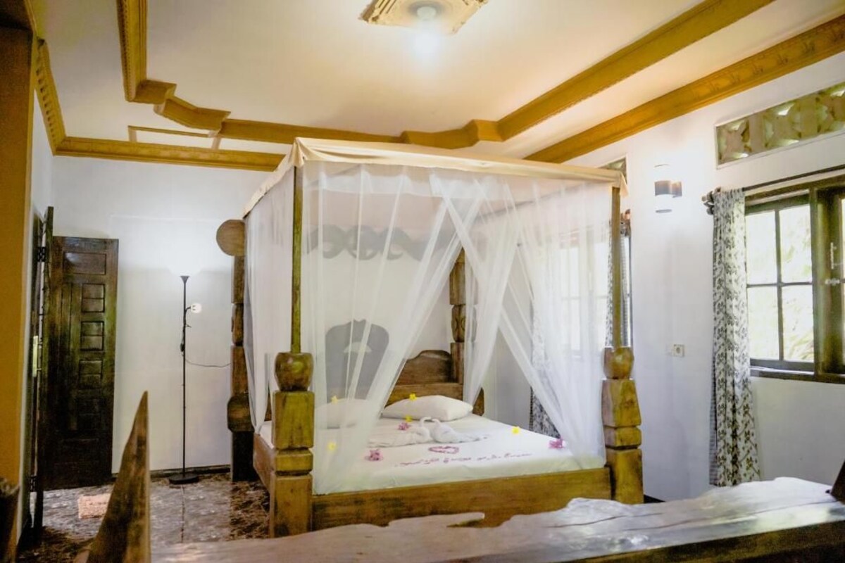 A four-poster bed with a canopy, wooden beams on the ceiling, a window with curtains, and a door with a wooden frame.