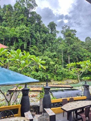 An outdoor seating area with tables and chairs, surrounded by lush greenery and a river.