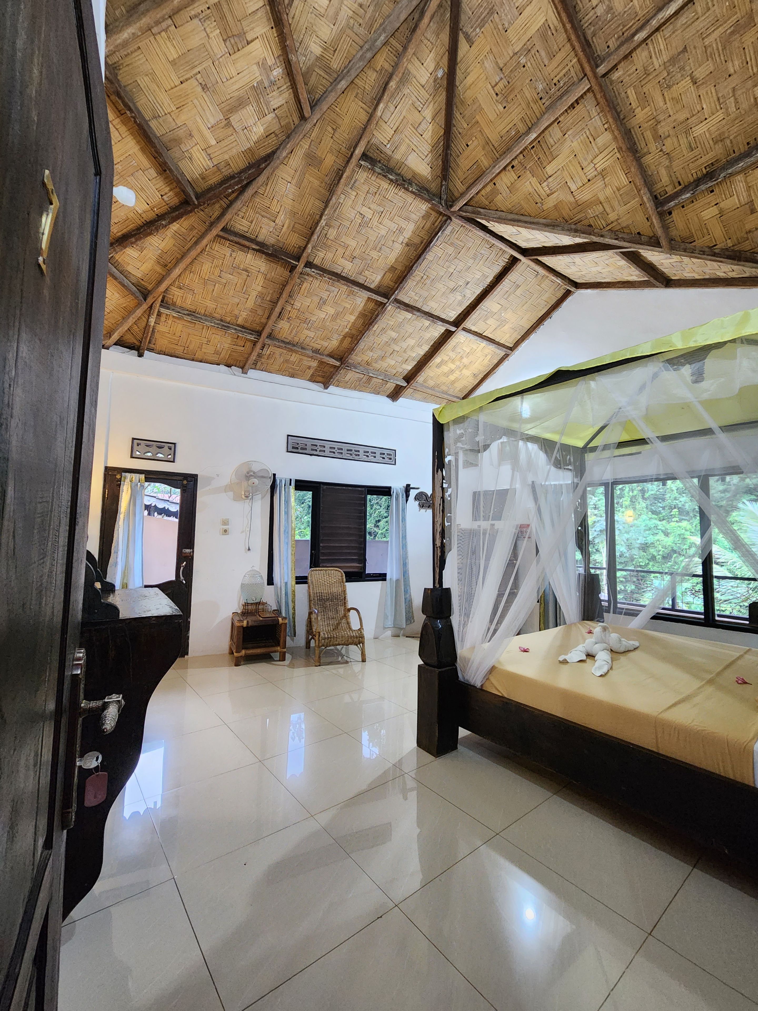 Una habitación con techo de paja, una cama con dosel, un ventilador y una silla.