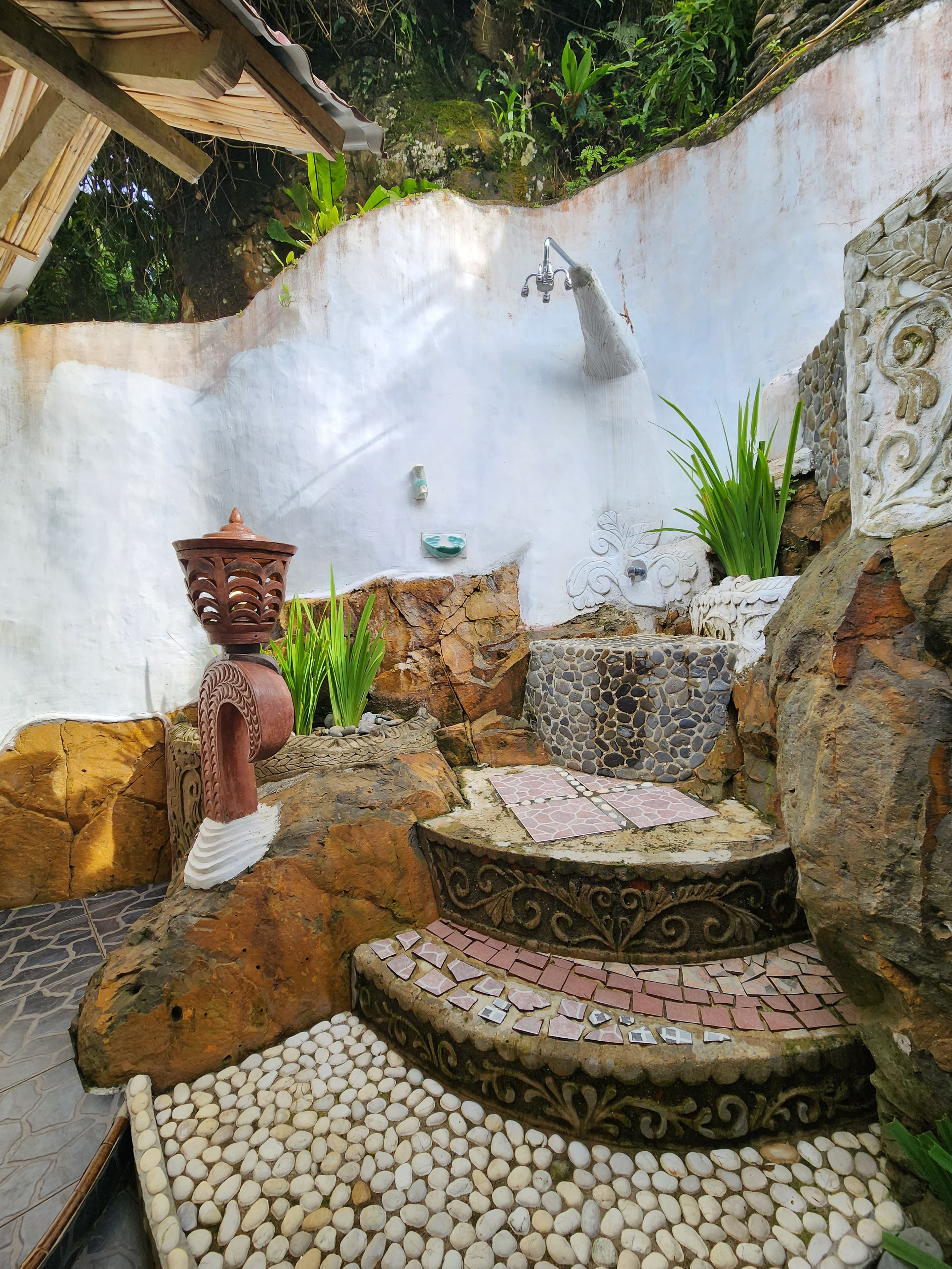 A stone-tiled patio with a decorative table, a rustic wall with a water spout, and a potted plant.