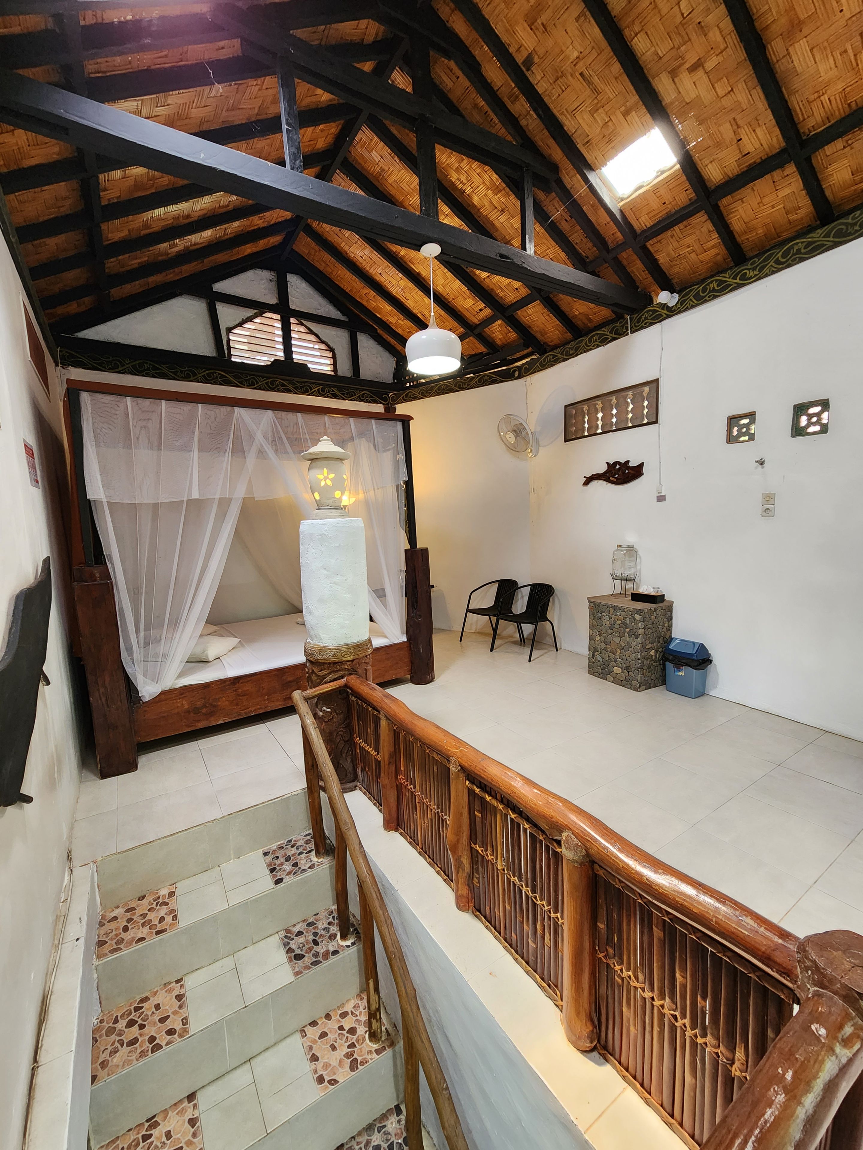 A room with a bed, a small table, a chair, a decorative wall piece, and a staircase with a wooden railing.