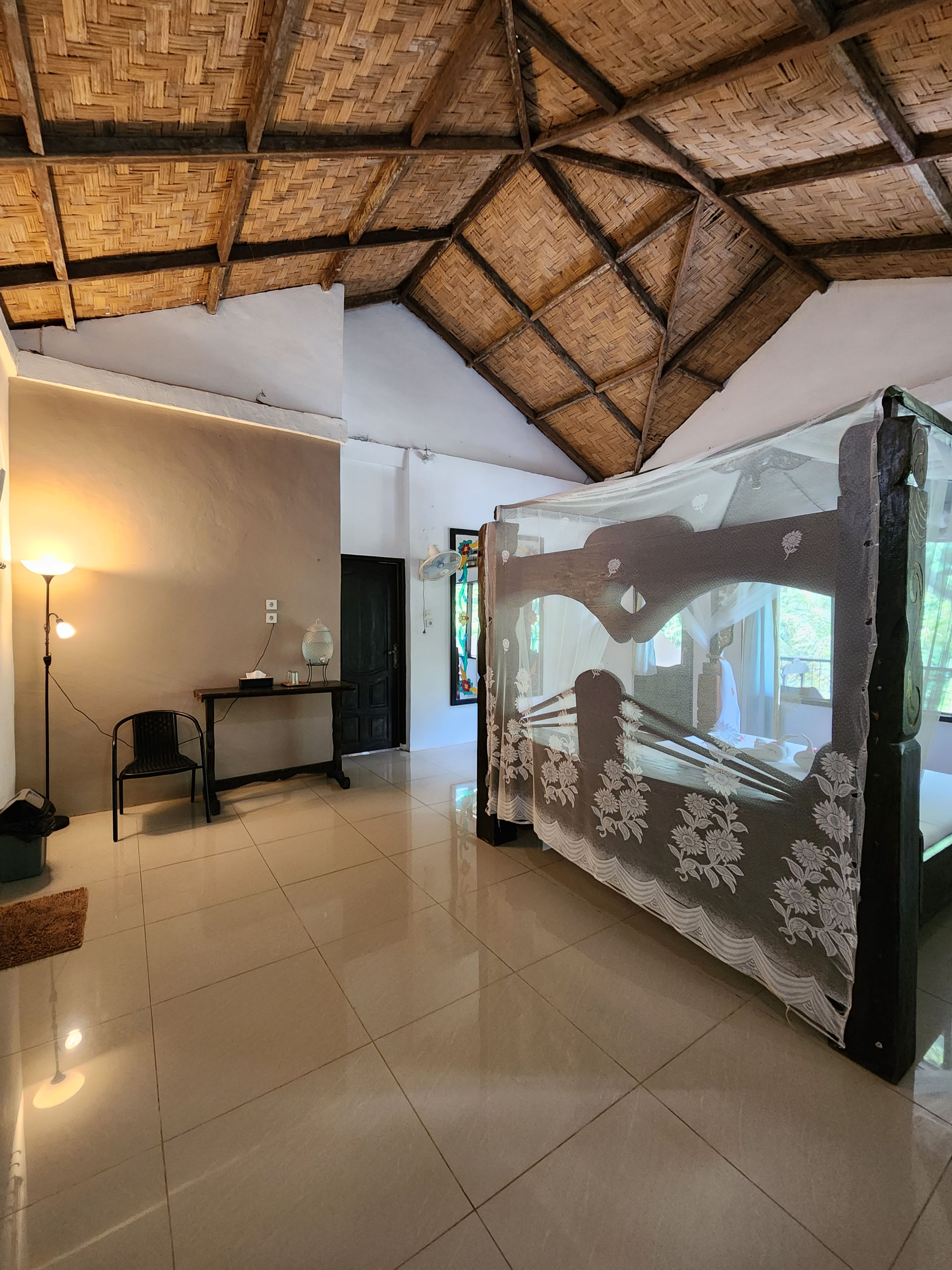 A room with a tiled floor, a bed with a floral bedspread, a wooden ceiling, a table with a lamp, and a chair.
