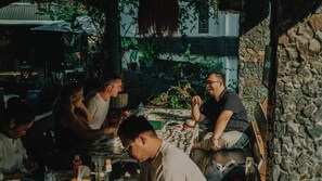 Un grupo de personas sentadas en una mesa al aire libre, con un muro de piedra y vegetación de fondo.