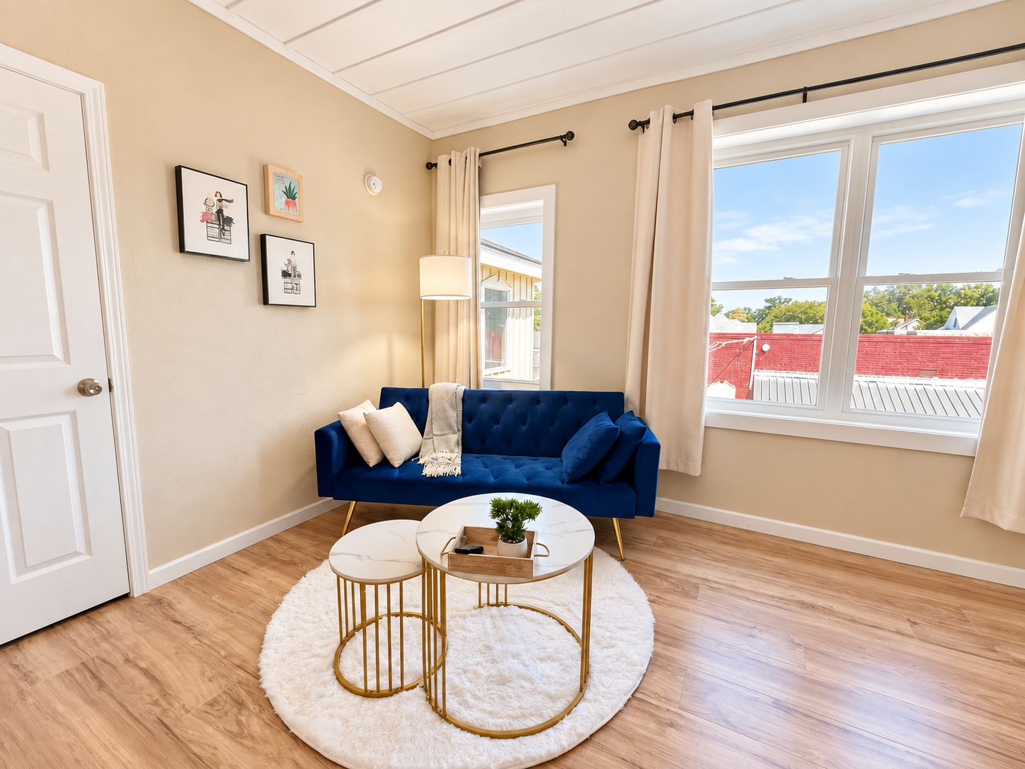 Een woonkamer met een blauwe bank, een ronde salontafel en een wit vloerkleed. Er hangen fotolijsten aan de muur, een lamp staat er en er is een raam met gordijnen.