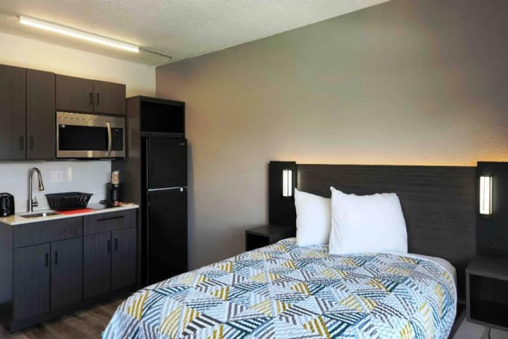 A hotel room with a bed, a nightstand, a headboard, a small kitchenette with a microwave, a refrigerator, and a sink.