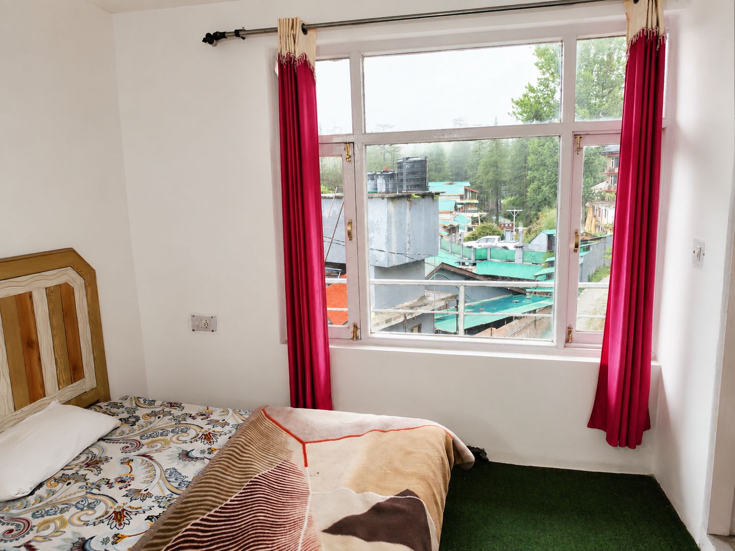 A bedroom with a bed, a window with curtains, and a view of buildings outside.
