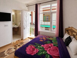 Un dormitorio con cama, ventana con cortinas, puerta que da a un baño y televisor en la pared.