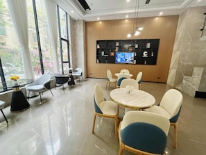 Un moderno vestíbulo de hotel con piso de mármol, mesas y sillas redondas, un televisor en la pared y grandes ventanales con cortinas.
