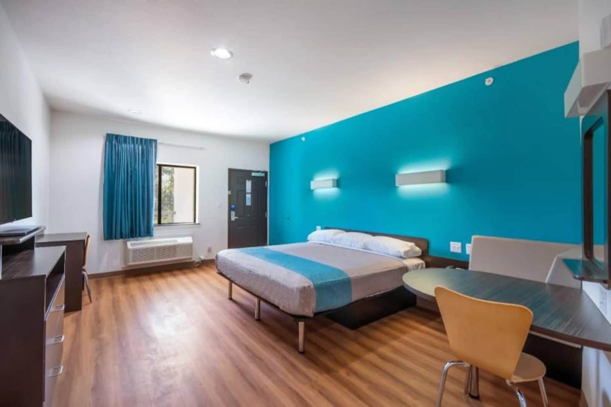 Habitació d'hotel amb llit, escriptori, cadira, finestra amb cortines i una paret decorativa blava.