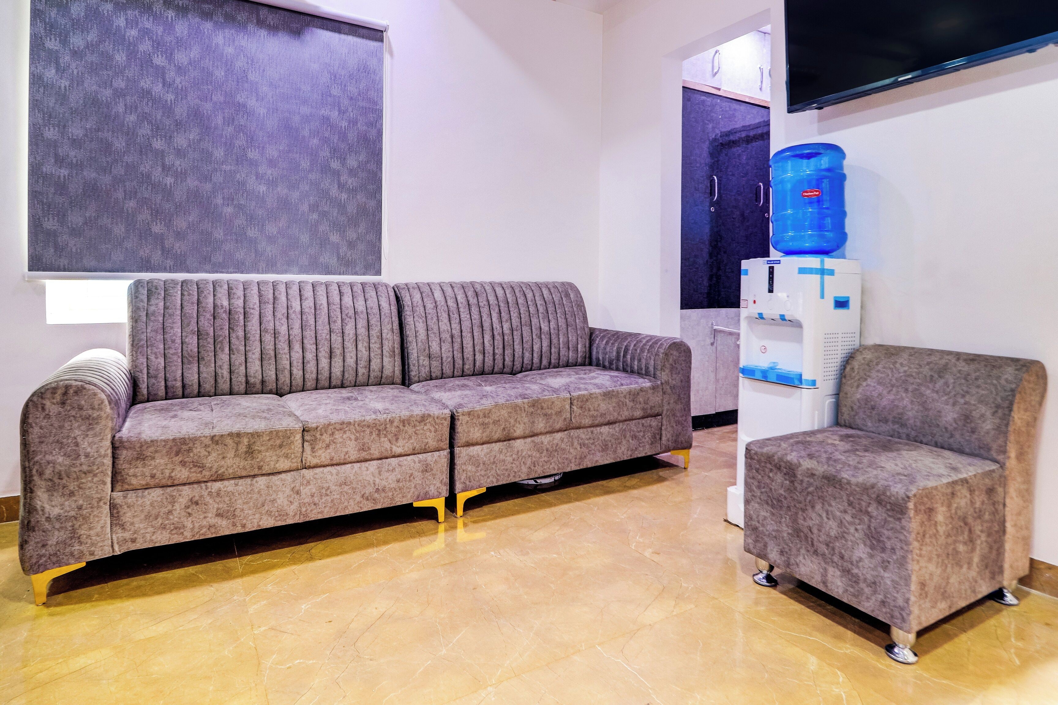 A modern living room with a grey sofa and a matching armchair, a water dispenser, and a flat-screen TV mounted on the wall.