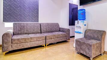 A modern living room with a grey sofa and a matching armchair, a water dispenser, and a flat-screen TV mounted on the wall.