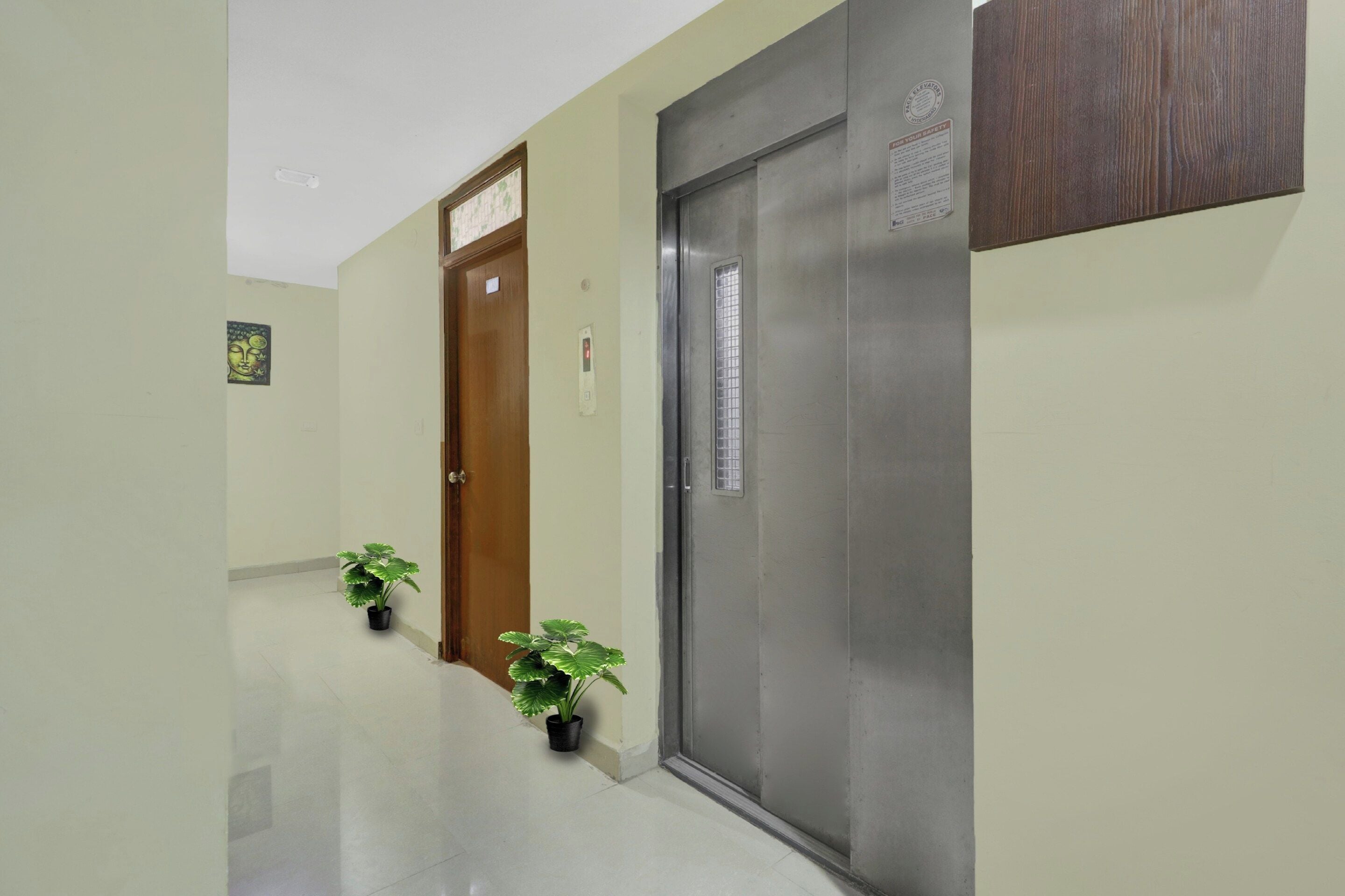 Un pasillo con una puerta de madera y una puerta de ascensor metálica, ambas con instrucciones de seguridad. Hay dos plantas en macetas en el suelo.