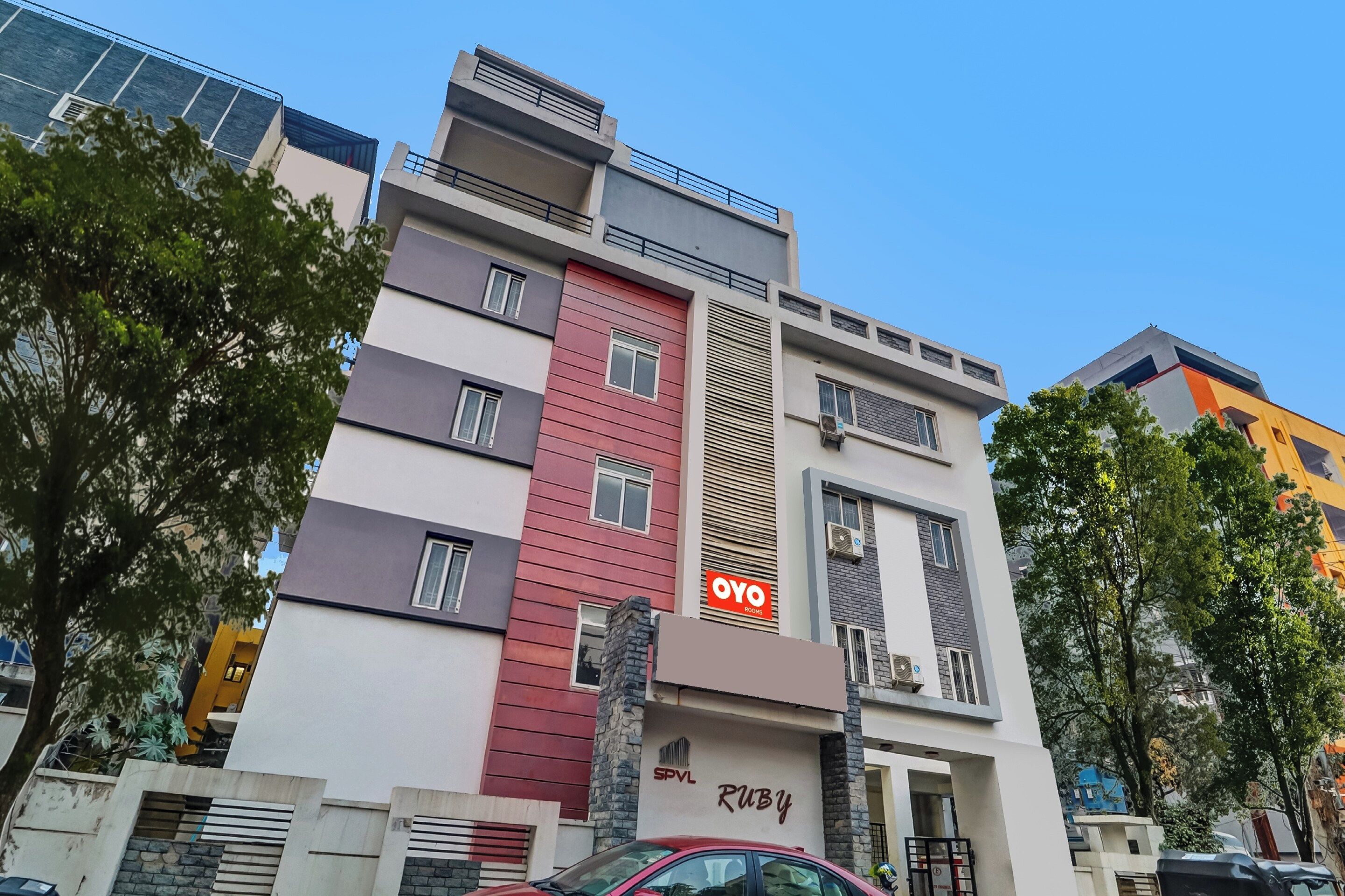 Un edificio de varios pisos con una sección vertical roja destacada, un letrero que dice 'OYO Rooms' y una entrada visible con un pilar de piedra.