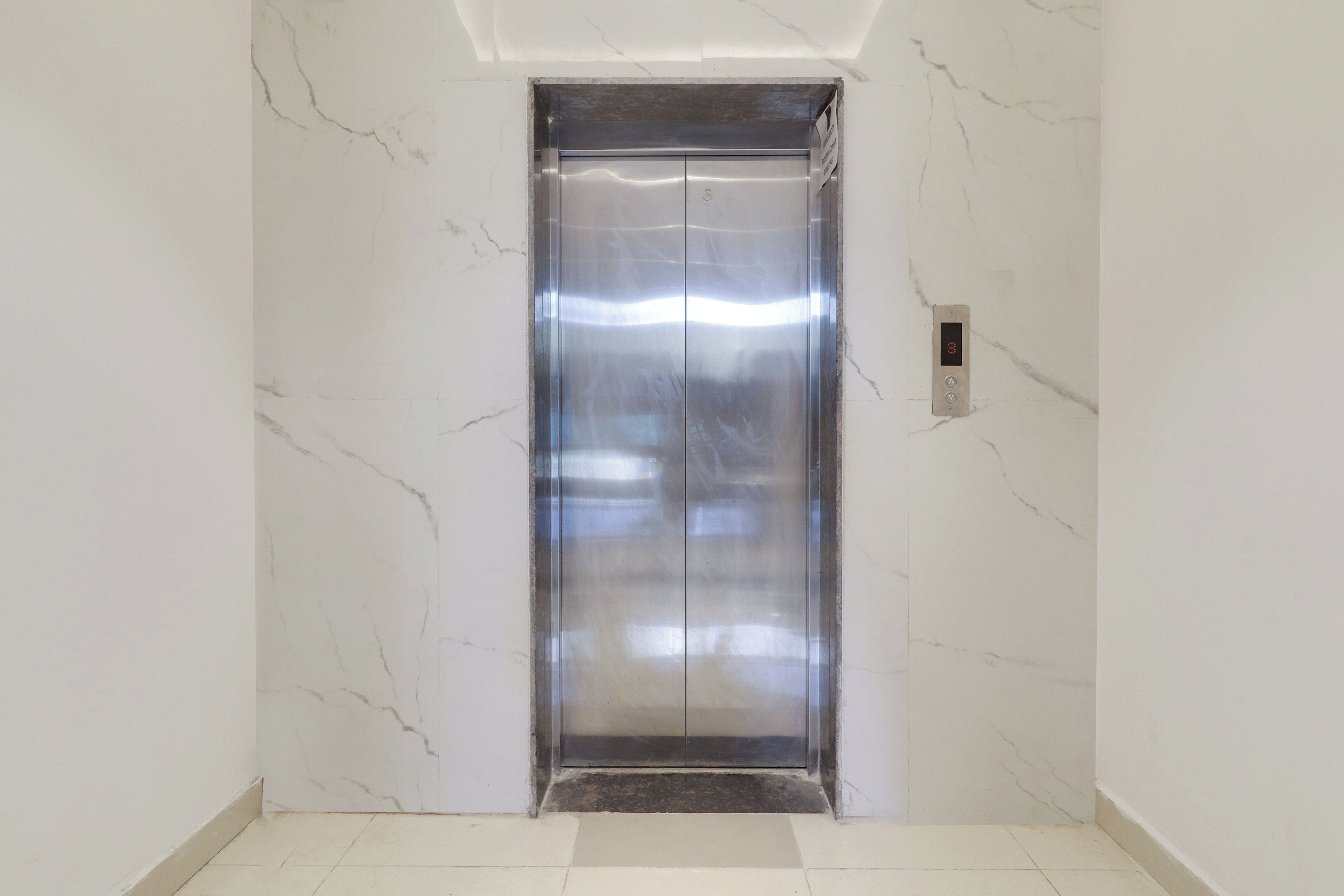 A modern elevator with a metallic finish, located in a hallway with white marble walls and floor tiles.