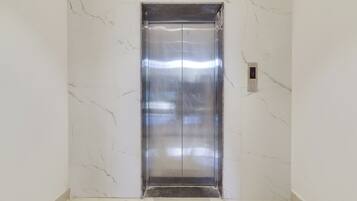 A modern elevator with a metallic finish, located in a hallway with white marble walls and floor tiles.