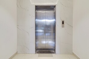 Un ascensor moderno con detalles metálicos, situado en un pasillo con paredes y suelo de mármol blanco.