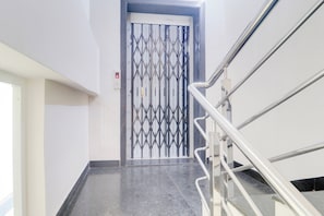 Un ascensor moderno con una puerta metálica geométrica, un pasamanos y un panel de control con una luz indicadora roja.