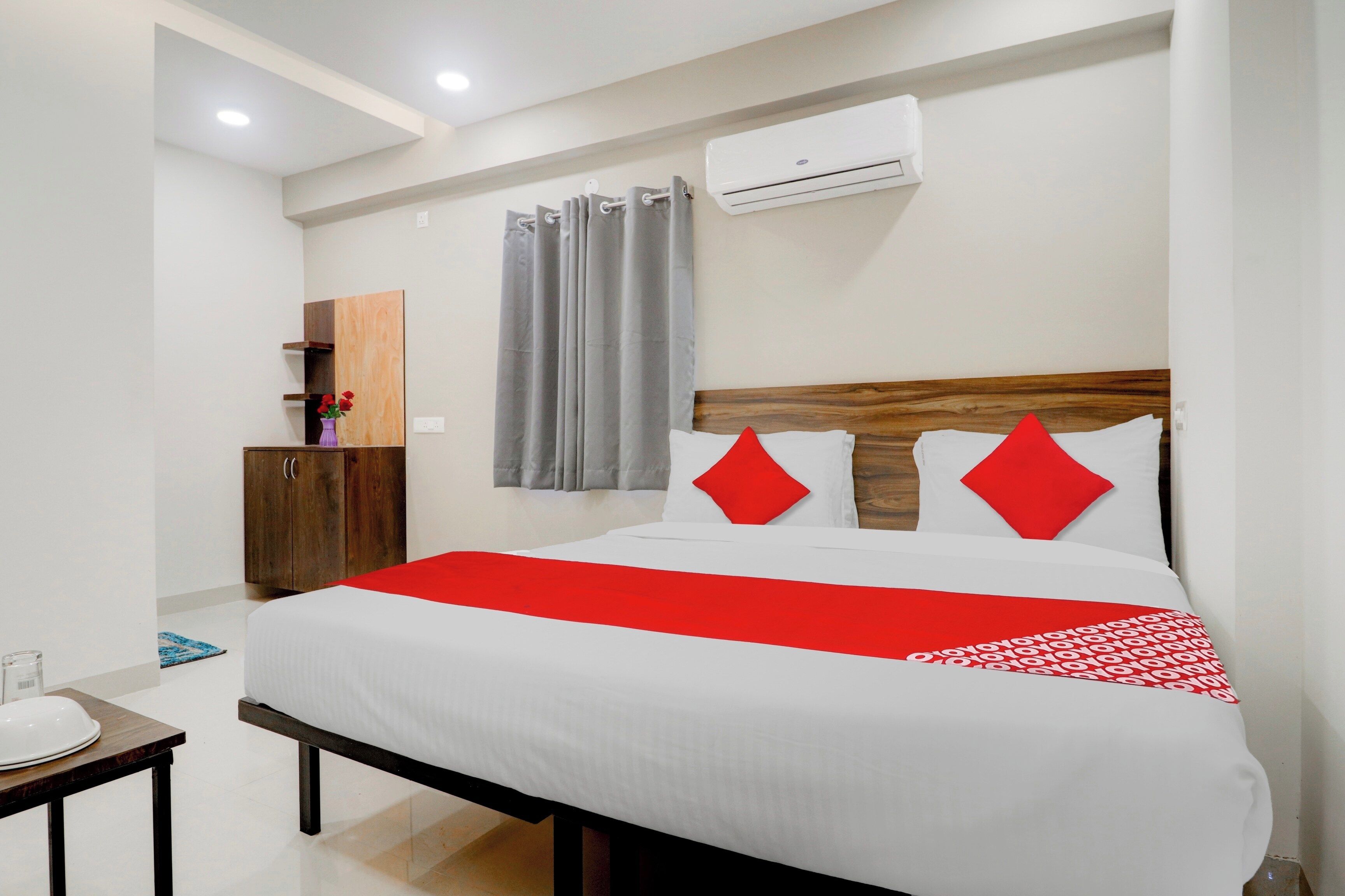 A hotel room with a bed, a wooden headboard, a red and white bedspread, a small table with a plate, and a wall-mounted air conditioner.
