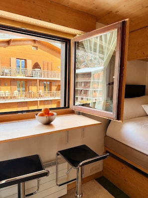 A compact room with a bed, a small table with a bowl of fruit, a window with curtains, and a view of a building with balconies. - New Listing (Verbier)