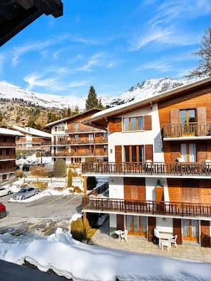 A multi-story hotel with balconies, a parking area, and snow-covered mountains in the background. - New Listing (Verbier)