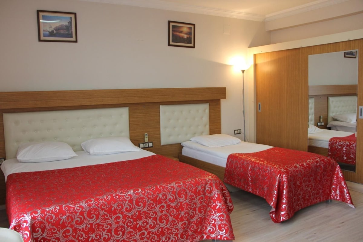 A hotel room with two beds, each with a red patterned bedspread and white pillows. There are two framed pictures on the wall, a bedside lamp, and a wooden headboard.