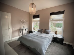 A bedroom with a large bed, bedside tables, a hanging light fixture, and a view of the outdoors through windows. - New Listing (Zeewolde)