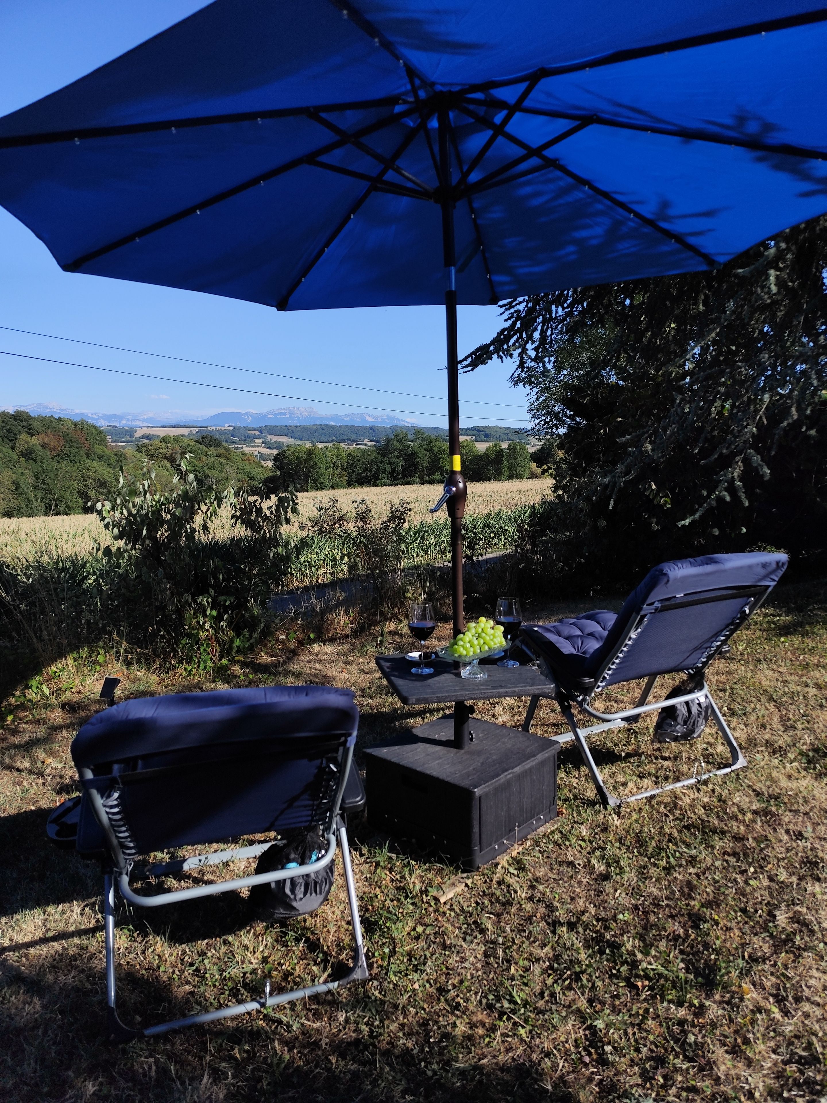 Ein Außenbereich mit Sitzgelegenheit, zwei Liegestühlen, einem Tisch mit einer Obstschale und einem großen, blauen Sonnenschirm, der Schatten spendet.