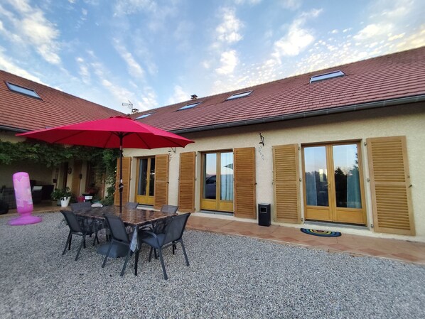 Un espace extérieur abrité, avec une table et des chaises, un parasol rouge et un sol en gravier.