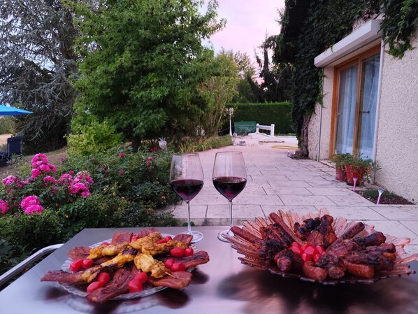 An outdoor dining area with a table set for two, featuring a variety of grilled meats and a glass of red wine.