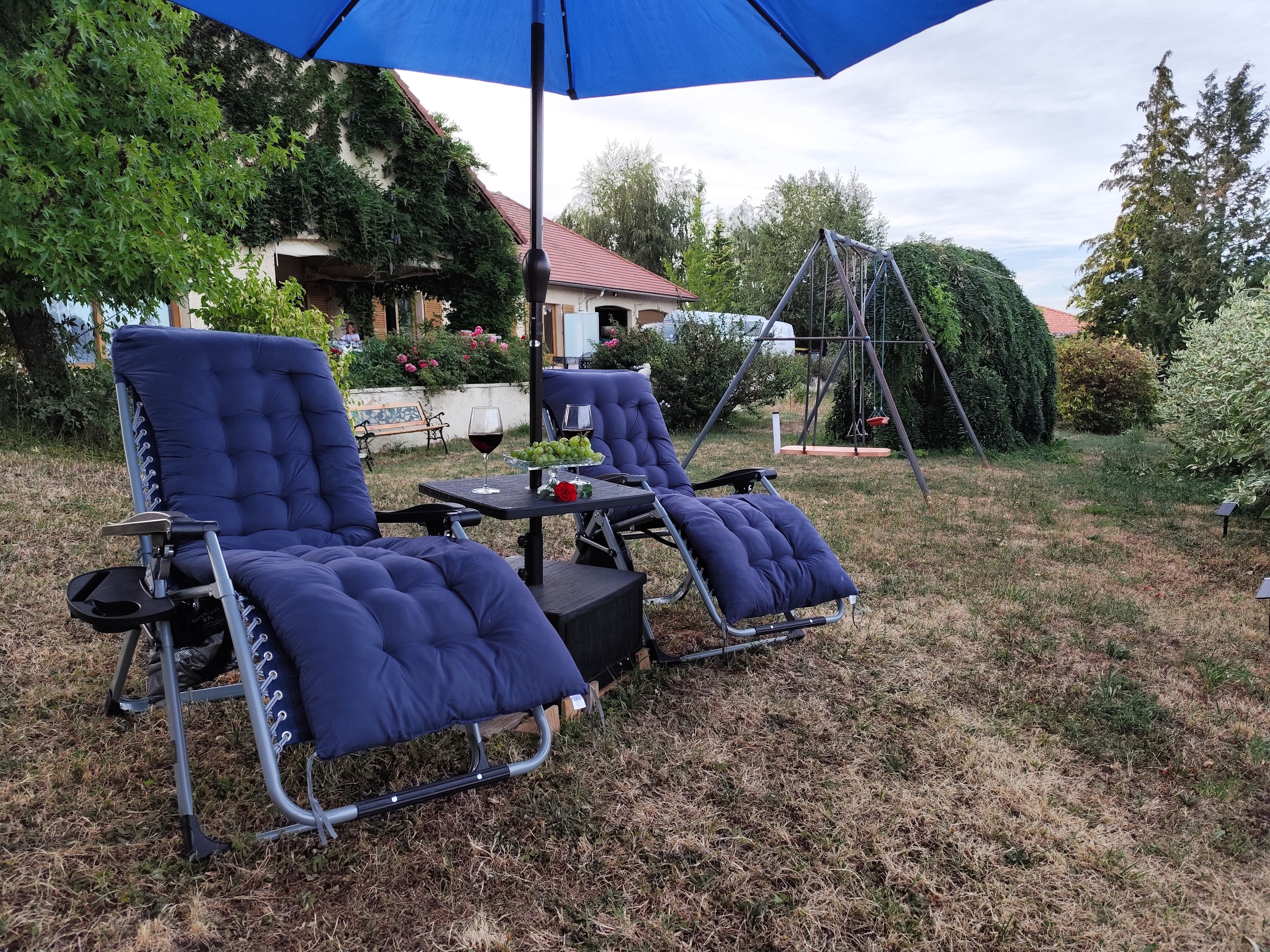 Two lounge chairs under a blue umbrella, a small table with a glass of red wine, and a swing set in a backyard.