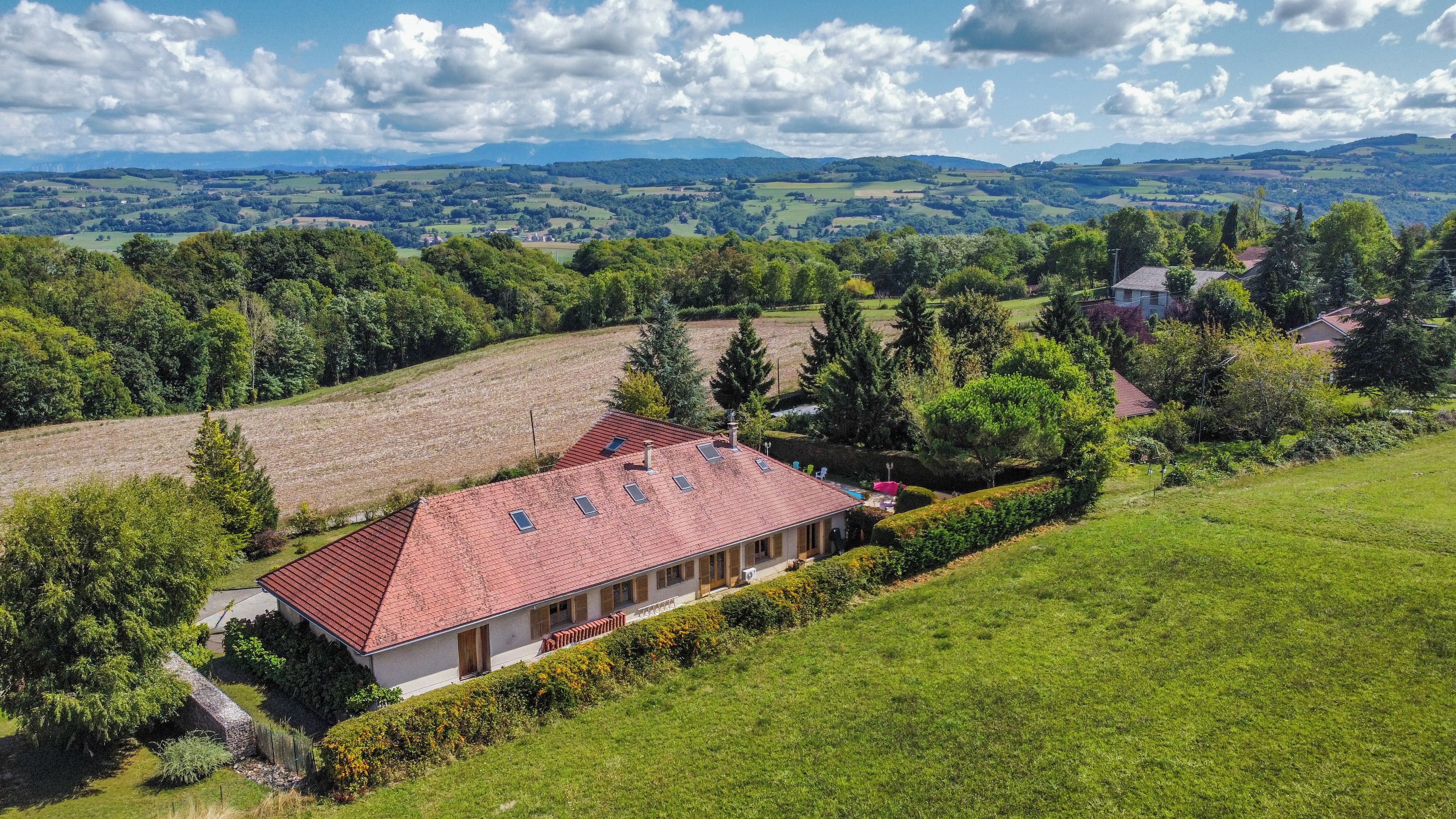 Une maison avec un toit rouge, entourée de champs verdoyants et d’arbres, offrant une vue sur des collines lointaines.