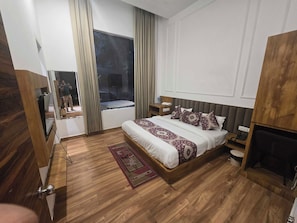 A hotel room with a large bed, a wooden floor, a window with curtains, a bedside table, and a television. - Hotel O Ak pride (Amritsar)