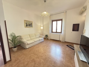 Un salon avec un canapé beige, une plante en pot, une lampe sur pied, une fenêtre avec des rideaux et un radiateur.