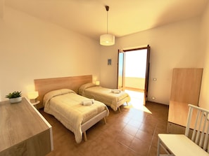 Une chambre d’hôtel avec deux lits, un bureau, une chaise, une plante en pot et une vue sur l’extérieur.