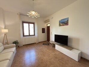 A living room with a sofa, a TV on a stand, a radiator, a window with curtains, and a ceiling light fixture. - Villa Horizon - Sky & Sea Terrace (Amalfi)