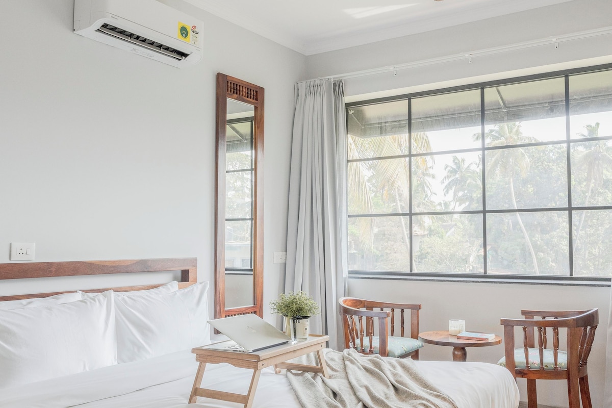 A bedroom with a bed, a desk, a chair, a ceiling fan, and a window with a view of palm trees.