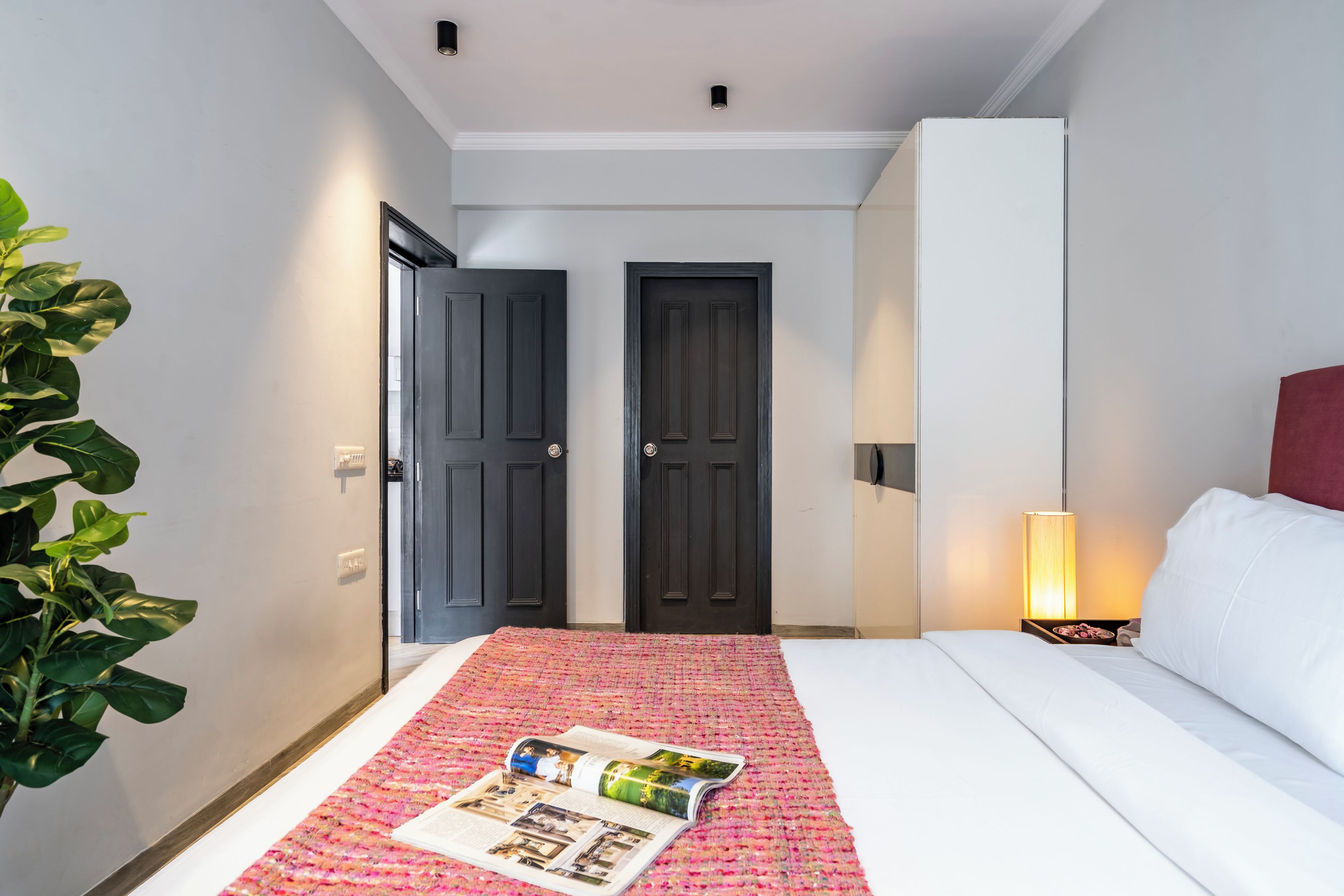 Habitación de hotel con cama, mesita de noche con lámpara, una puerta y una planta.