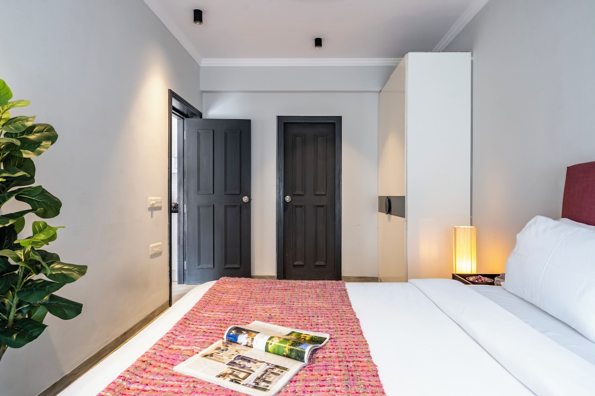 A hotel room with a bed, a bedside table with a lamp, a door, and a plant.