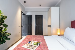 A hotel room with a bed, a bedside table with a lamp, a door, and a plant. - Staymaster Avtara - Serviced Apartments (Calangute)