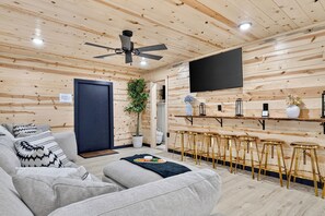 Ein modernes Interieur mit Holzbalkendecke, Deckenventilator, Flachbildfernseher, Bar mit Hockern und einer Couch mit gemusterten Kissen.