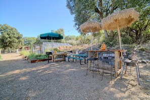 Zona d'estar a l'exterior amb taula i cadires, un para-sol verd i una estructura de fusta rústica.