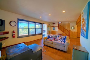 A spacious living area with a large window, a sofa with a sports-themed blanket, a fire extinguisher, and a wooden staircase. - Spacious Beautiful Country Home on 8 Private Acres (Kelseyville)