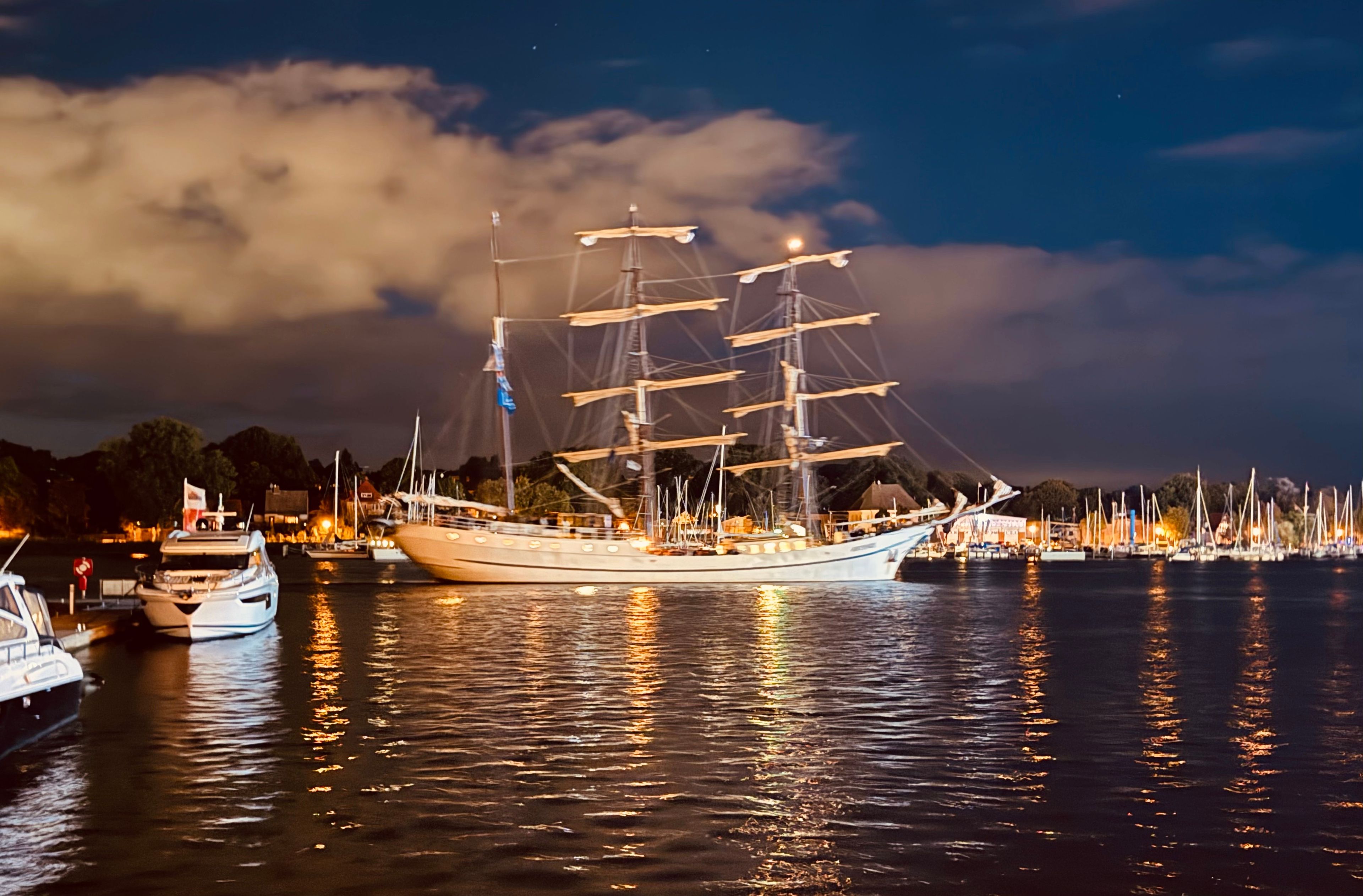 Ein großes Segelschiff liegt in einer Marina vor Anker, mit weiteren Booten und einer Stadtkulisse im Hintergrund.