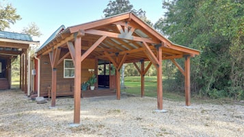 Un gazebo in legno con tetto spiovente, sorretto da travi di legno, su una superficie ghiaiosa con un'area erbosa e alberi sullo sfondo.