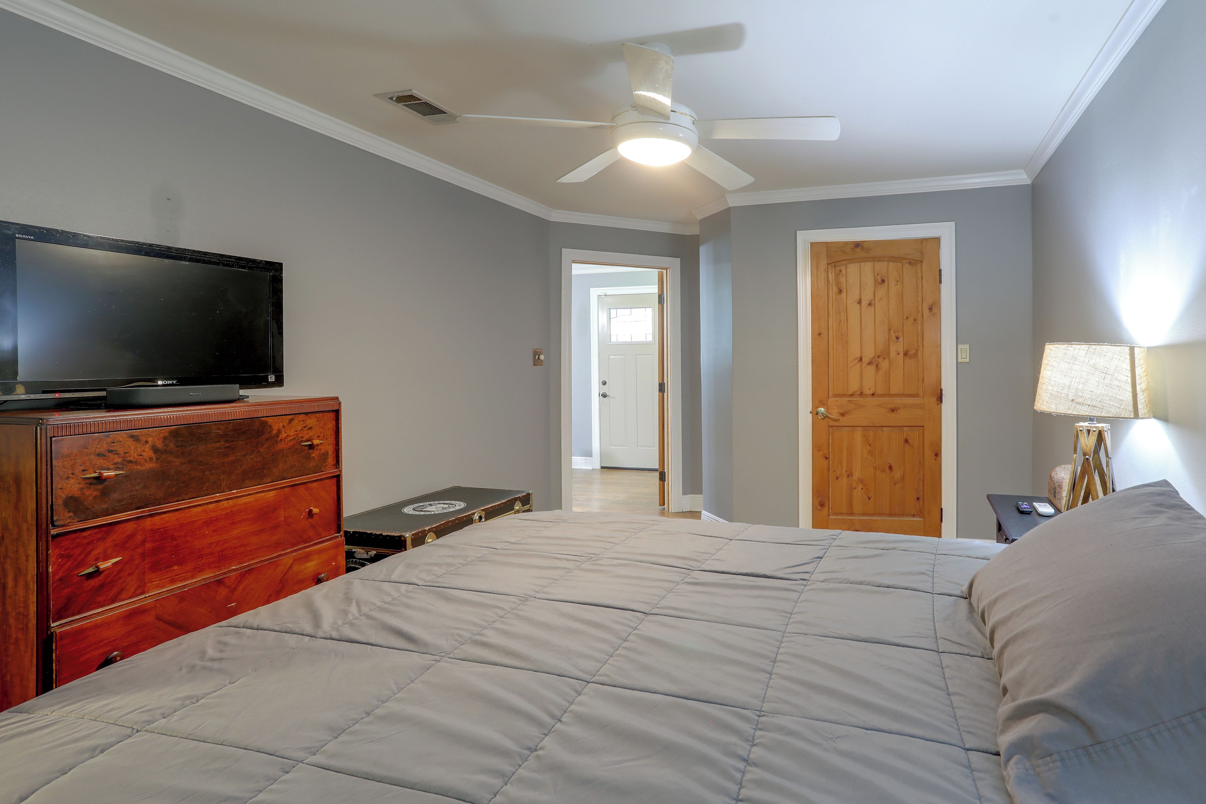 A bedroom with a bed, dresser, television, ceiling fan, and a door leading to another room.