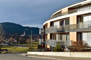 Ein modernes Gebäude mit einer gewölbten Fassade, Balkonen mit Metallgeländern und Blick auf eine Stadt sowie Berge im Hintergrund.