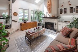 A spacious living room with a fireplace, a sofa, a coffee table, and a view of the outdoors through large windows. - Luxury Manhattan Beach Home in Parklike Gated Community (Manhattan Beach)