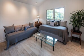 A modern bedroom with a glass coffee table, two beds with matching bedding, a nightstand with a lamp, and a window with a view of trees. - Luxury Manhattan Beach Home in Parklike Gated Community (Manhattan Beach)