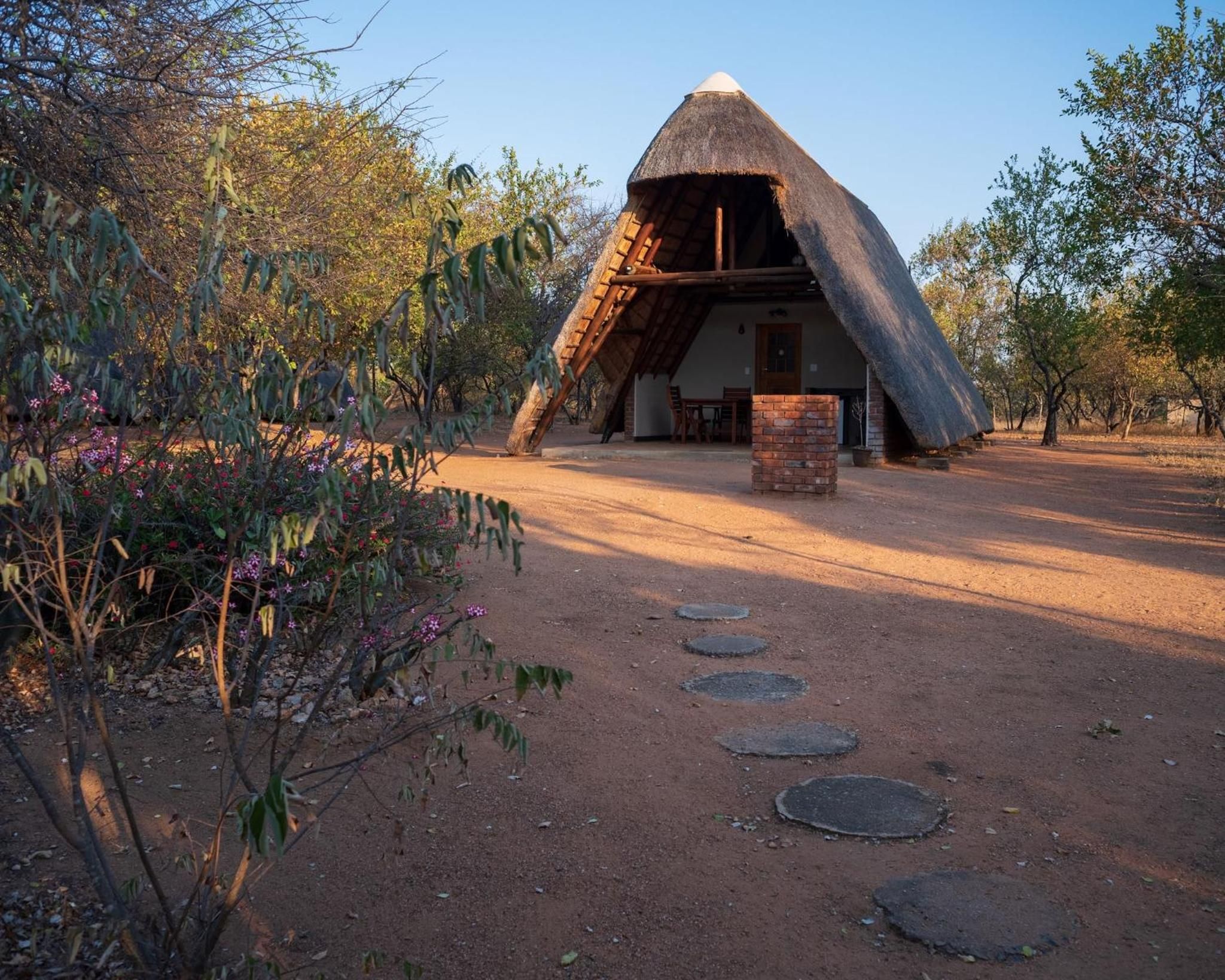 Un bâtiment à toit de chaume, doté de fondations en briques, entouré d’un chemin de terre et de végétation.